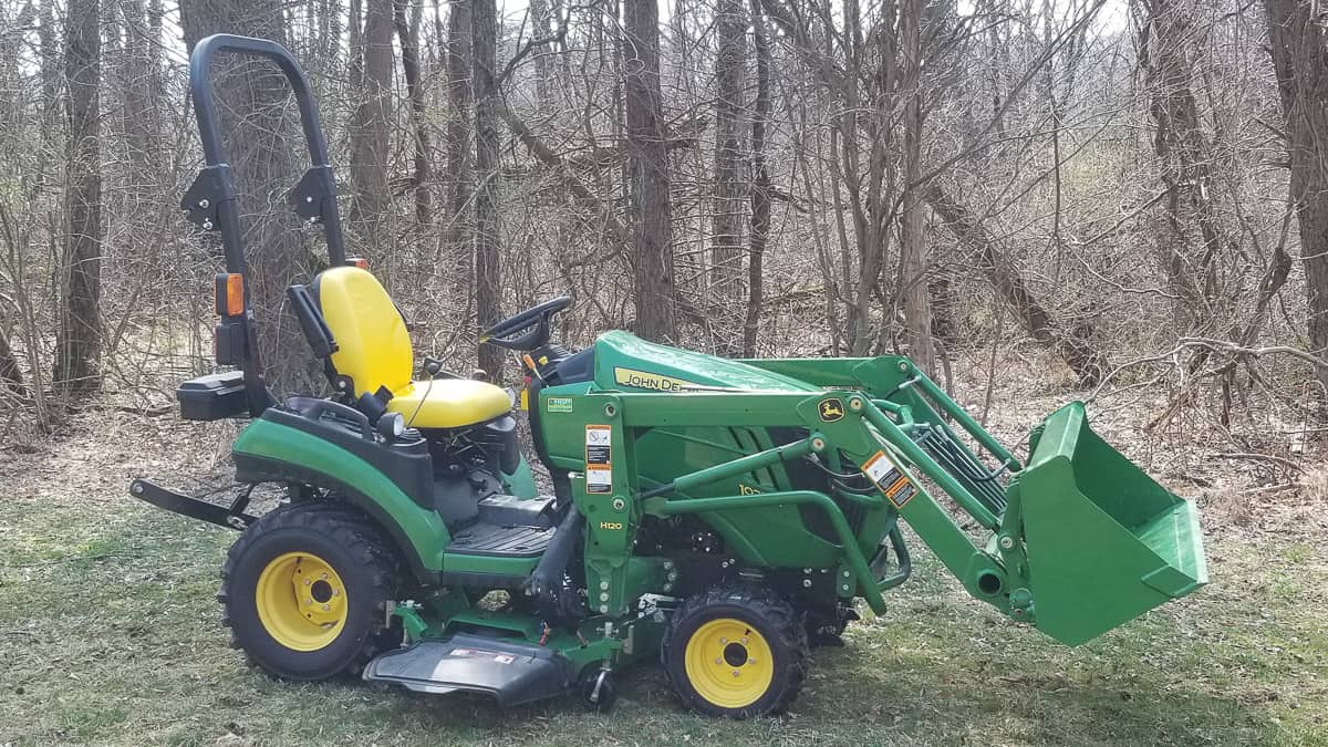 A green John Deere sub-compact utility tractor with a front loader bucket and mowing deck, parked in a wooded area.