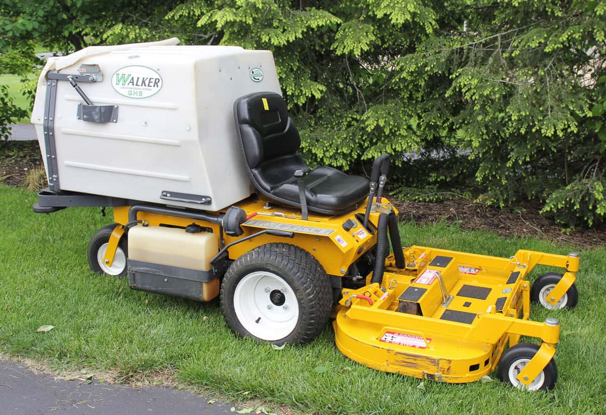 A yellow and white Walker brand riding lawn mower parked on grass near bushes.