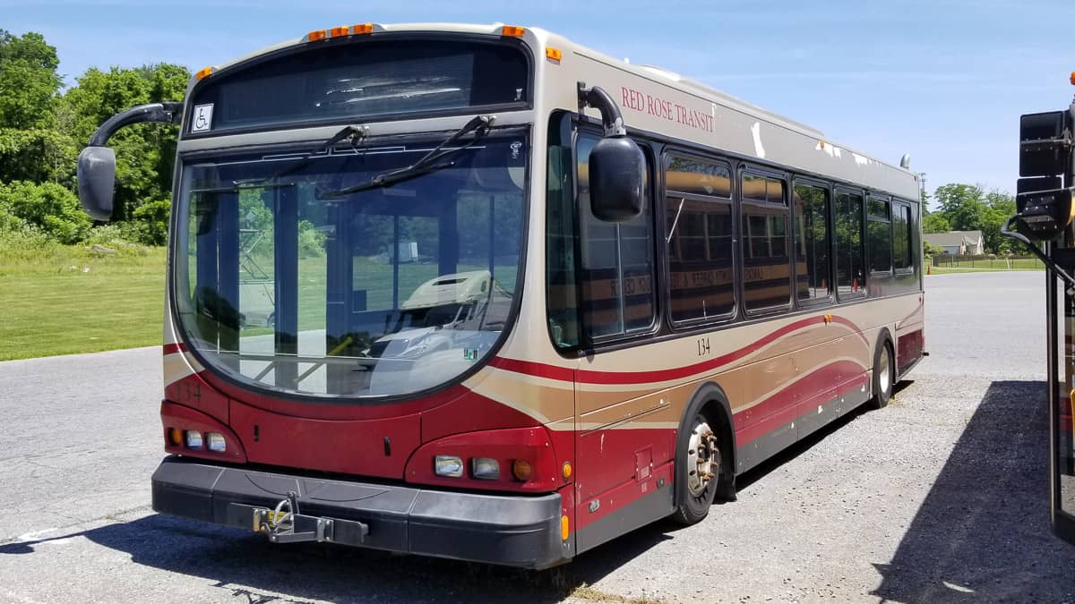 Red and beige public transit bus parked in a sunny open lot with clear blue skies above.