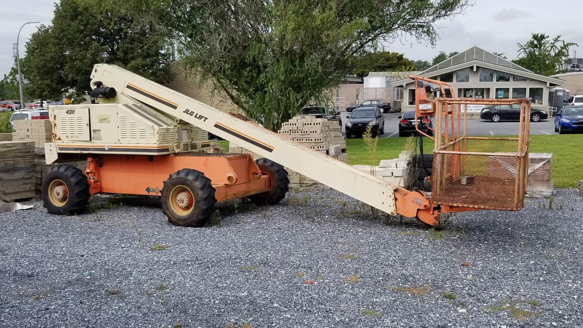 An orange and beige telescopic boom lift parked on a gravel lot near pallets of bricks and a green lawn.