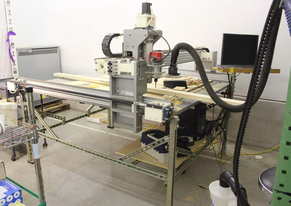 Industrial workshop with a CNC machine, computer setup, and various tools on a cluttered table, inside a brightly lit room.