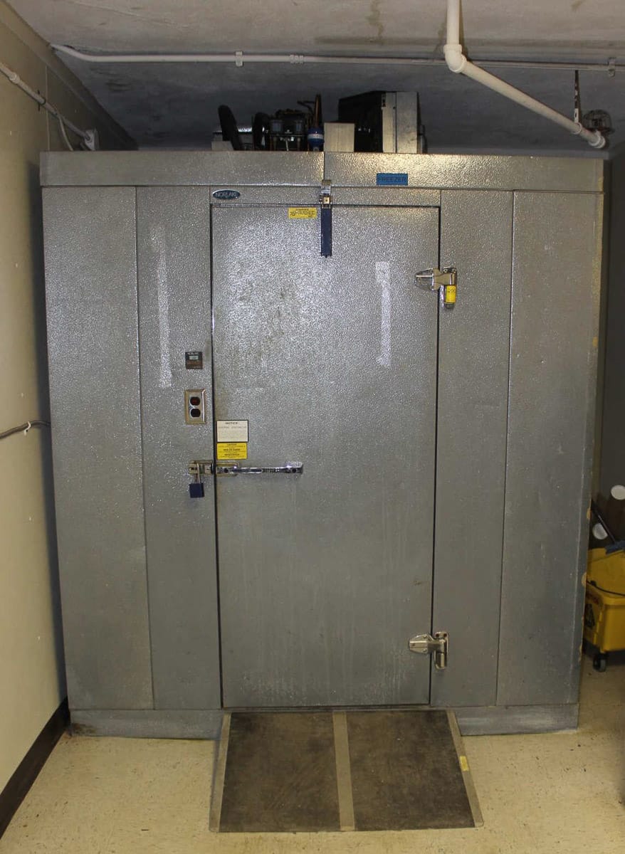 Large gray industrial electrical cabinet with multiple locks and warning labels, in a dimly lit room with concrete flooring.