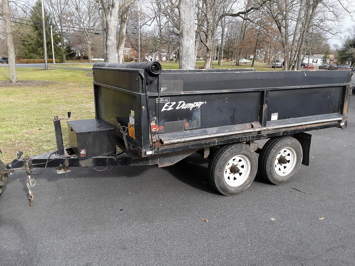 A black EZ Dumper utility trailer attached to a tow hitch, parked on an asphalt surface with grass and trees in the background.