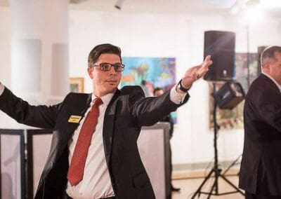 A man in a suit and glasses gestures with open arms at a fundraiser, with another man partially visible in the background.