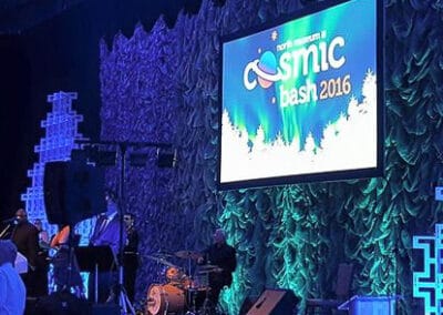 A band about to perform onstage at the "Cosmic Bash 2016" event, with a large blue-lit screen displaying the event title.