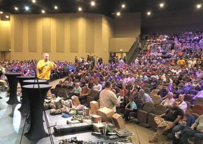 A man in a yellow shirt speaking at a podium to a large audience in an auditorium, with various hunting gear displayed on stage.