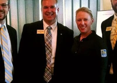 Three professionals, two men and one woman, smiling at an event, wearing name tags and formal attire.