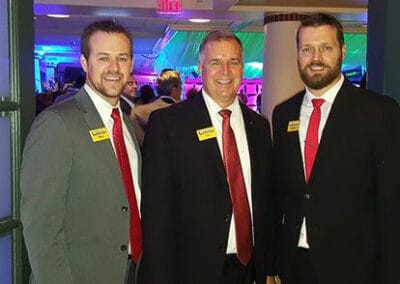 Three smiling men in suits with name tags standing in a room with colored lighting.