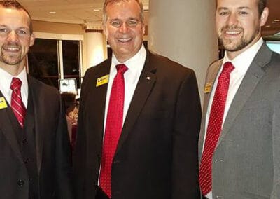 Three smiling men in business suits with red ties standing together at an indoor event.
