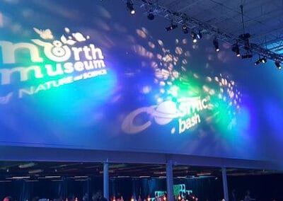 Event venue with people at tables under blue lighting and projected logos for "North Museum" and "Cosmic Bash" on the wall.