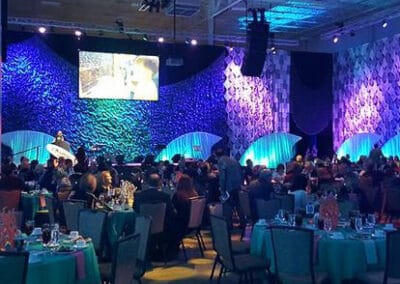 Elegant room with guests seated at tables, a speaker on stage, and large screen displaying an event, under blue lighting.