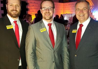 Three smiling men in business suits with matching red ties and name tags standing together at an event.