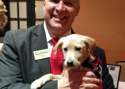 Man in a suit smiling and holding a light brown puppy wearing a black and red scarf.