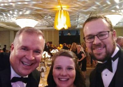 Three people smiling for a selfie at a formal event with a chandelier overhead and blurred guests in the background.