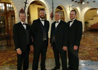Four men in tuxedos smiling in a grand hall with vintage decor and arches.
