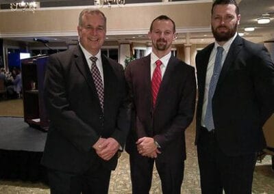 Three men in suits smiling at a formal event, standing in a carpeted room with a stage in the background.