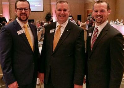 Three smiling men in suits, two wearing yellow ties and one in a striped tie, standing at a formal event.