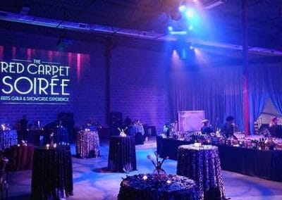 Event venue illuminated in blue lighting, with round tables and a sign saying "The Red Carpet Soiree" on the brick wall.