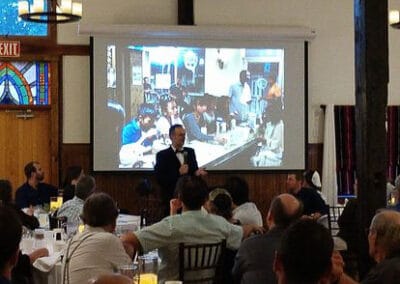 A man speaks at a podium in a room with attendees listening and a large screen displaying a video conference in the background.