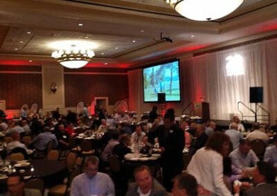 People at a formal event with round tables, in a large hall with a stage and a projected presentation in the background.