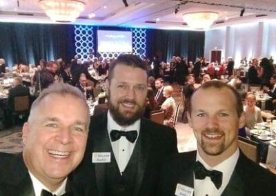 Three smiling men in tuxedos at a fundraising event with a crowded banquet hall in the background.