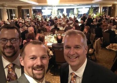 Three men smiling at a busy gala dinner in an elegantly decorated ballroom with chandeliers and seated guests.