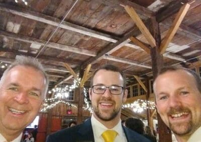 Three men in suits smiling at a rustic indoor event, adorned with fairy lights on wooden beams above.