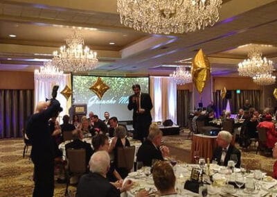 Elegant room with guests seated at tables, a speaker on stage, and large chandeliers overhead.