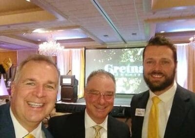 Three smiling men in suits at an event, with name tags and a background screen displaying the word "Gretna".