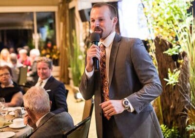 Man in a suit speaking into a microphone at a formal event with seated guests listening in the background.