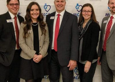 Five people, three men and two women, smiling and posing at a professional event in front of a banner with the MCAP logo.