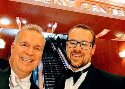 Two smiling men in tuxedos inside a lobby with a lit-up sign saying "Event Center" and an escalator in the background.