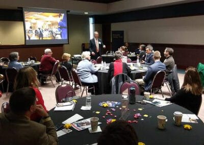 A speaker presents to a group of attendees inside a well-lit room.