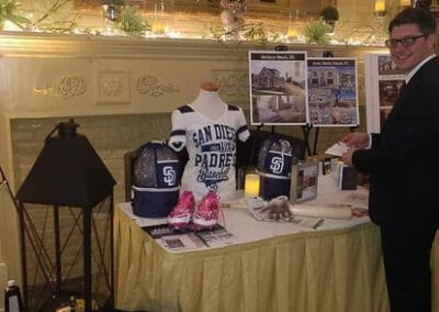 A man in a suit stands at a display table with San Diego Padres merchandise, informational posters, and brochures.