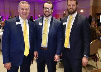 Three men in suits with matching yellow ties smiling at a formal event, standing in a banquet hall.