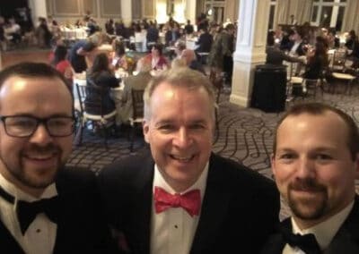 Three men in tuxedos smiling for a selfie at a formal event with guests seated at round tables in the background.