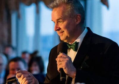 A middle-aged man in a tuxedo with a green bow tie speaks into a microphone at a fundraising event.
