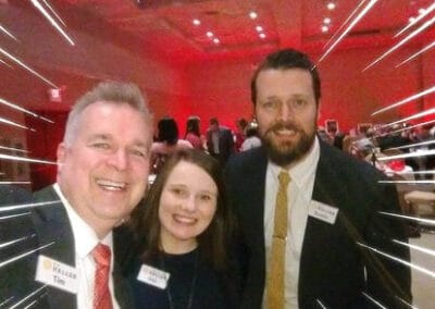 Three people wearing name tags smiling at a fundraising event, with bright white light streaks radiating around them.