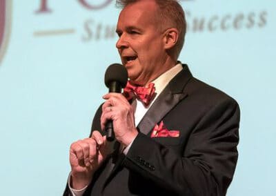 Man in a tuxedo with a red bow tie speaking into a microphone on stage, with a banner in the background.