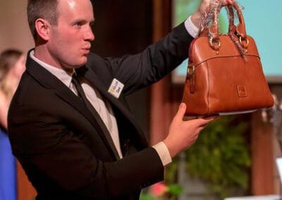A man in a suit presenting a brown leather handbag at an auction.