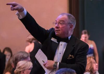 An auctioneer in a tuxedo energetically pointing and speaking at an event, with attendees in the background.
