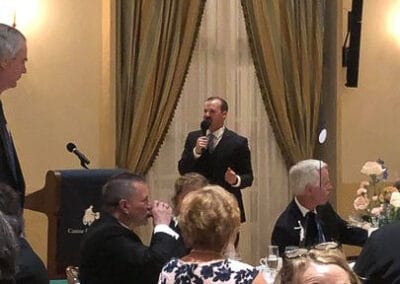 A man speaks at a podium to an audience during a fundraiser in a room with elegant curtains and festive balloons.