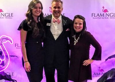 Three people posing with smiles at a charity event, standing in front of a banner with flamingo motifs and event logos.