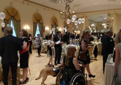 Guests, including a person in a wheelchair with a service dog, mingle in an elegantly decorated banquet hall during a fundraiser.