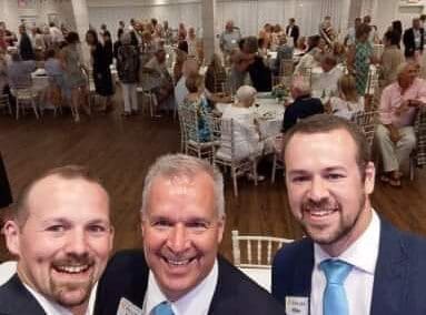 Three smiling men in suits taking a selfie at a busy fundraising event with many guests seated and standing in the background.