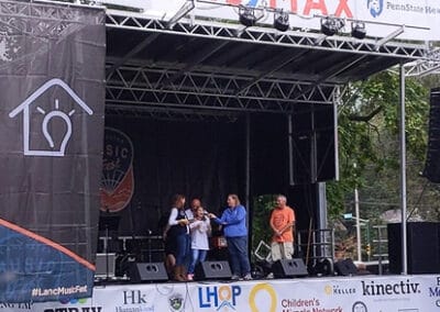 An outdoor event with five people on a stage sponsored by Re/Max, with company banners visible around the platform.