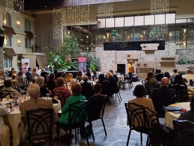 A room with attendees at round tables, listening to a speaker at a podium, with a lush indoor garden and hanging light fixtures.