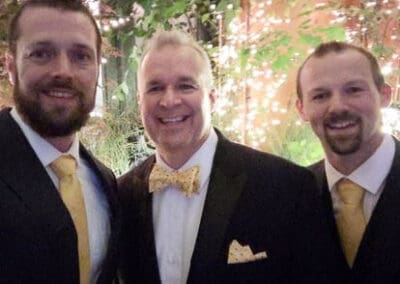 Three men in suits, two with yellow ties and one with a yellow bow tie, smiling during an evening event with a lit backdrop.
