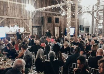 People at a formal event in a rustic barn decorated with fairy lights, listening to a speaker at the front.