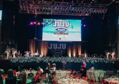 An event with decorated tables and attendants standing around, and a large screen displaying the logo "JUJU" onstage.